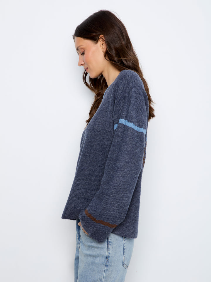 A woman wearing a dark heathered blue knit cardigan with light blue and brown stripe details on the sleeves, paired with light-wash jeans, photographed against a plain white studio background.