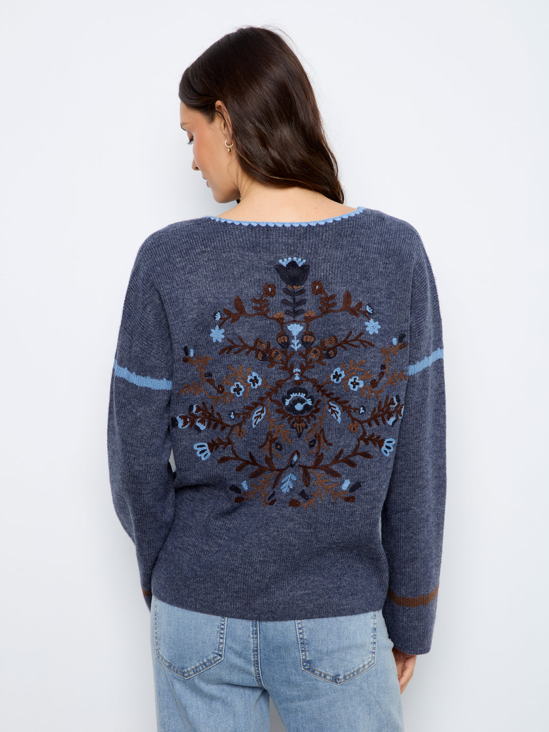A woman viewed from the back wearing a heathered slate blue knit sweater with a large brown and light blue floral pattern across the back, paired with light blue jeans against a plain white background.
