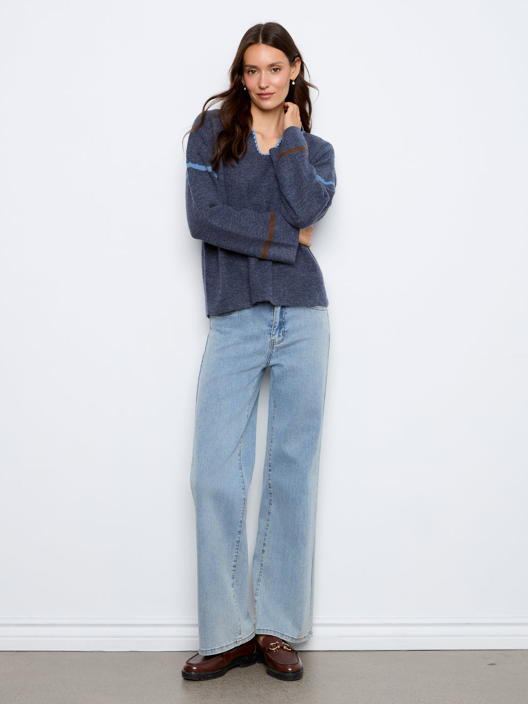 A woman stands against a white wall wearing a dark blue-gray knit sweater with light blue and brown stripe details on the sleeves, light-wash wide-leg jeans, and dark brown loafers.