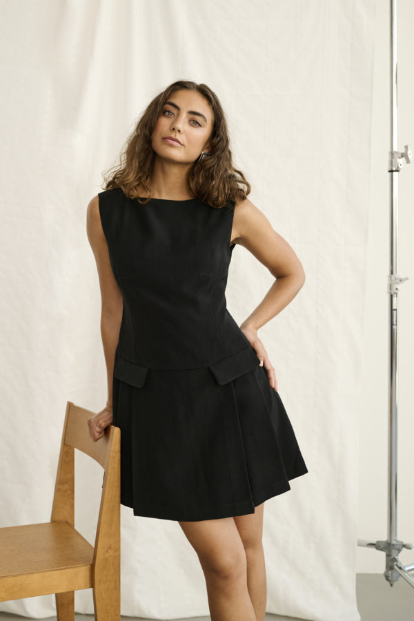Woman wearing a black dress standing next to a wooden chair against a white curtain.