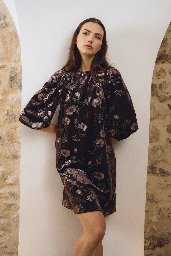 A woman models a short puff-sleeve dress made of dark purple velvet with a muted floral pattern, standing in a white arched alcove flanked by stone walls.