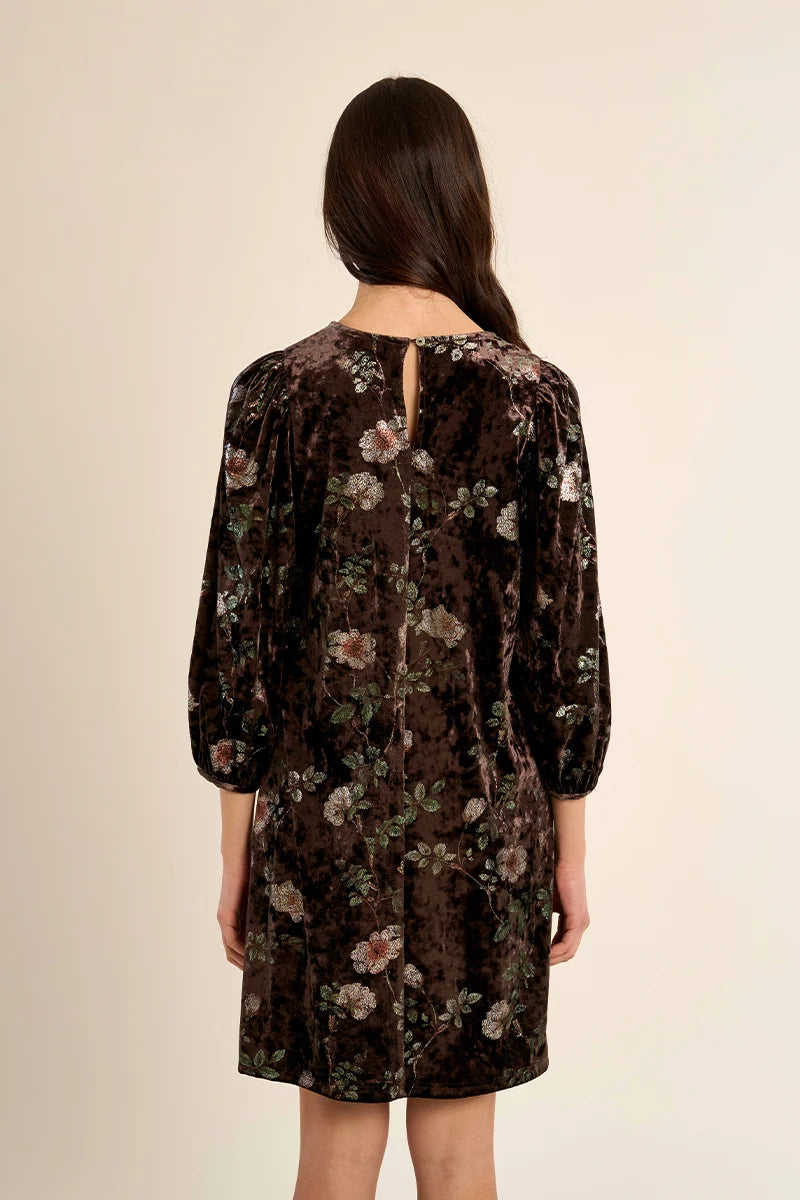 A woman viewed from the back wearing a dark brown velvet dress with a floral pattern, featuring three-quarter sleeves and a small keyhole cutout at the neckline, set against a plain beige background.