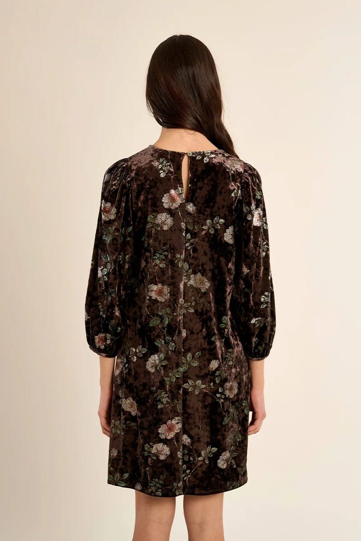 A woman viewed from the back wearing a dark brown velvet dress with a floral pattern, featuring three-quarter sleeves and a small keyhole cutout at the neckline, set against a plain beige background.