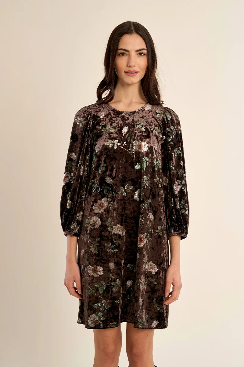 A woman modeling a dark brown velvet dress with a muted floral pattern in pink, green, and beige, set against a plain beige studio background.