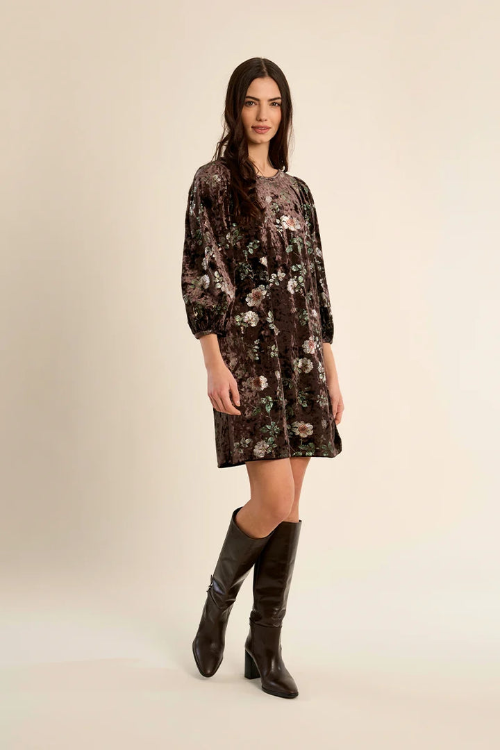 A woman modeling a dark brown velvet dress with a floral pattern, paired with black leather knee-high boots, against a solid light beige studio background.