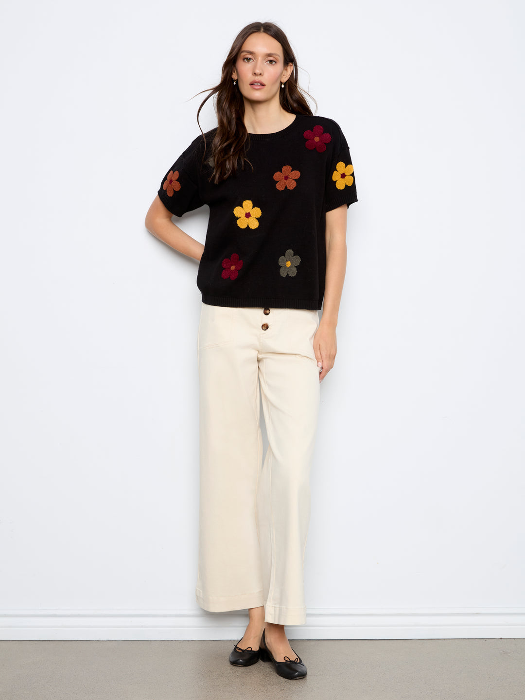 A woman stands against a white wall wearing a black short-sleeve knit top with colorful embroidered flowers and cream wide-leg cropped pants with black flats.