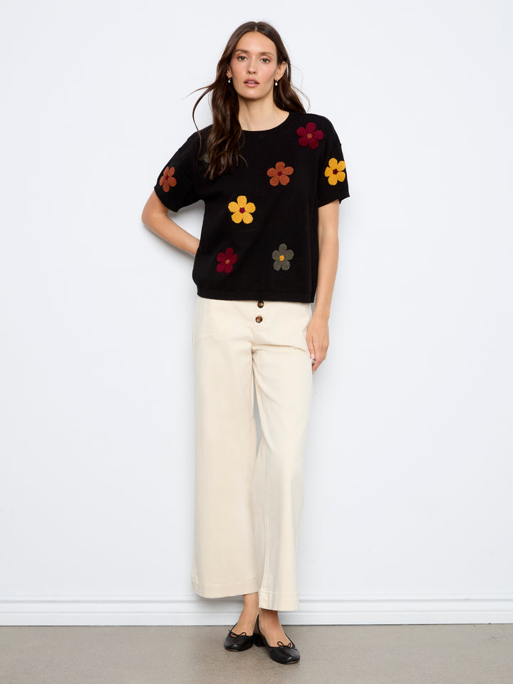 A woman stands against a white wall wearing a black short-sleeve knit top with colorful embroidered flowers and cream wide-leg cropped pants with black flats.