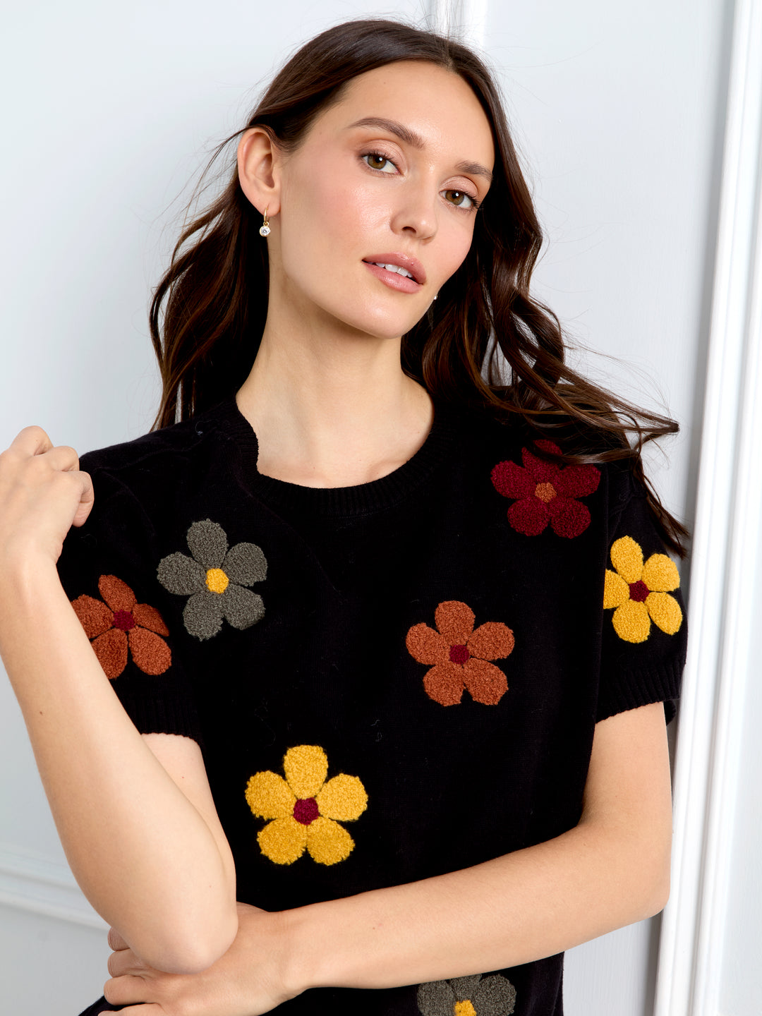 A woman wearing a black short-sleeve knit top with colorful knitted flower motifs in yellow, brown, and red, posed against a light gray interior background.