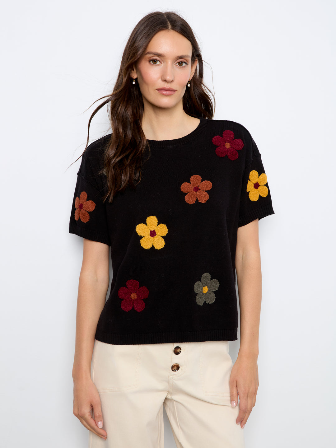 A woman modeling a black short-sleeve knit top with colorful embroidered flowers in yellow, red, brown, and gray, paired with cream pants against a light gray studio background.