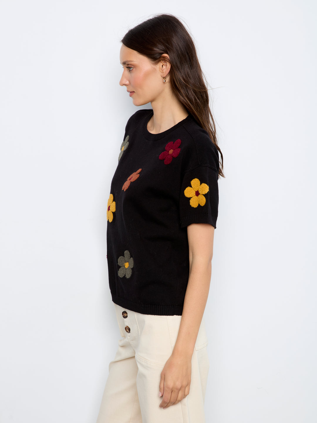 A woman wearing a black short-sleeve knit sweater with colorful embroidered flowers, paired with cream pants, photographed against a plain white studio background.