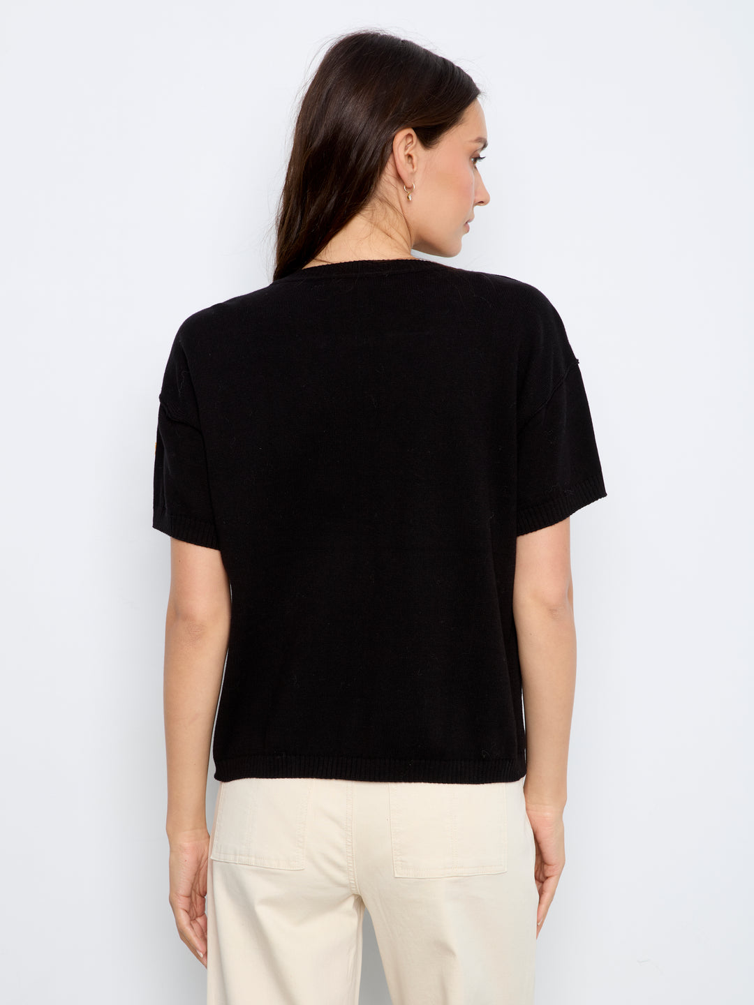 A woman shown from the back wearing a black short-sleeve crewneck top and cream-colored pants, photographed against a plain light gray studio background.