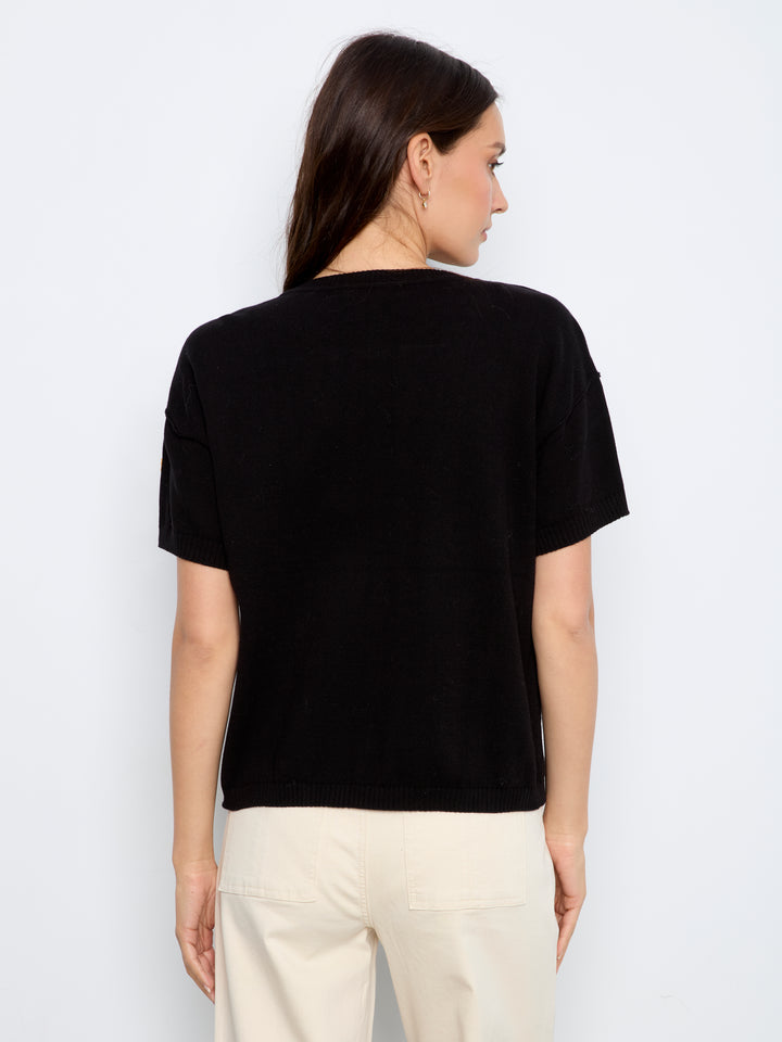 A woman shown from the back wearing a black short-sleeve crewneck top and cream-colored pants, photographed against a plain light gray studio background.