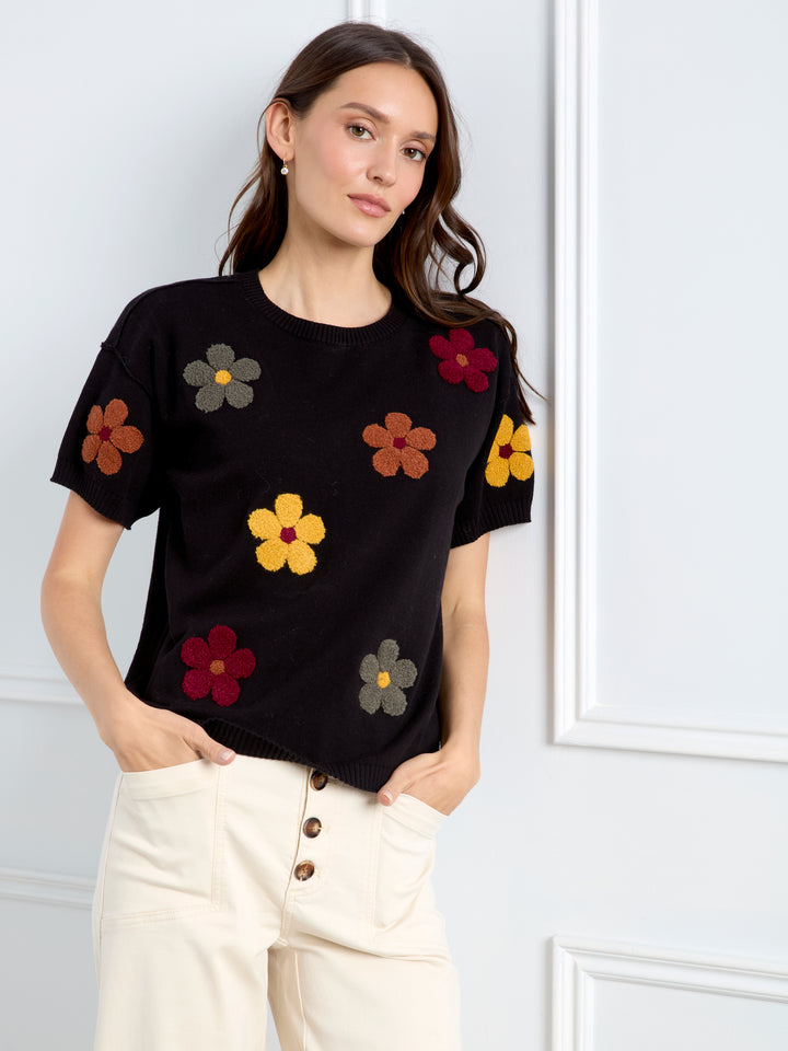 A woman wearing a black short-sleeve knit top with colorful embroidered flowers in yellow, red, brown, and gray, paired with cream-colored pants, standing against a white paneled wall.