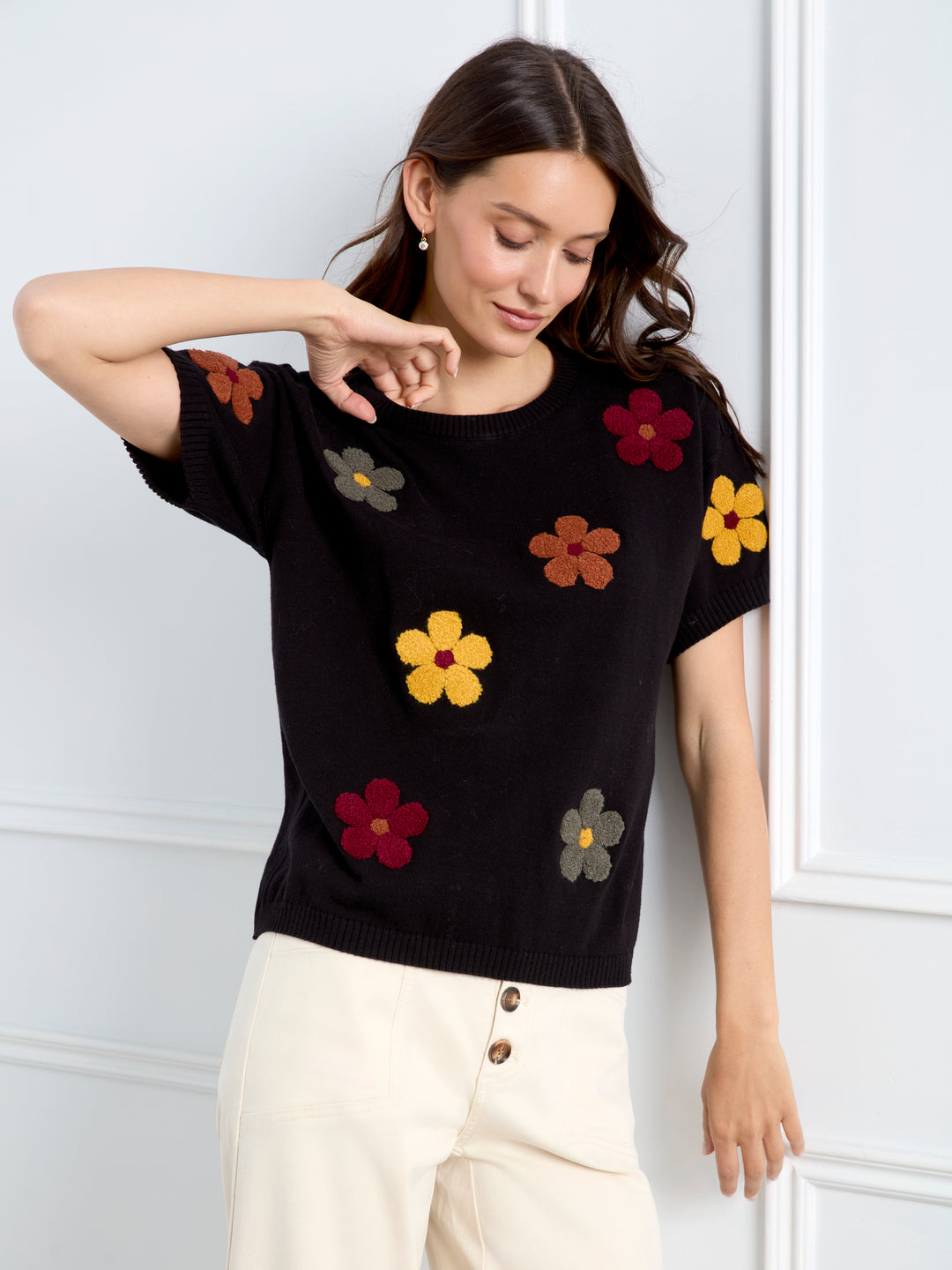 A woman wearing a black short-sleeve knit top with colorful embroidered flowers in yellow, red, brown, and gray, paired with cream-colored pants, posing against a white paneled wall.