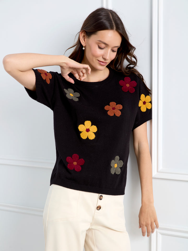 A woman wearing a black short-sleeve knit top with colorful embroidered flowers in yellow, red, brown, and gray, paired with cream-colored pants, posing against a white paneled wall.