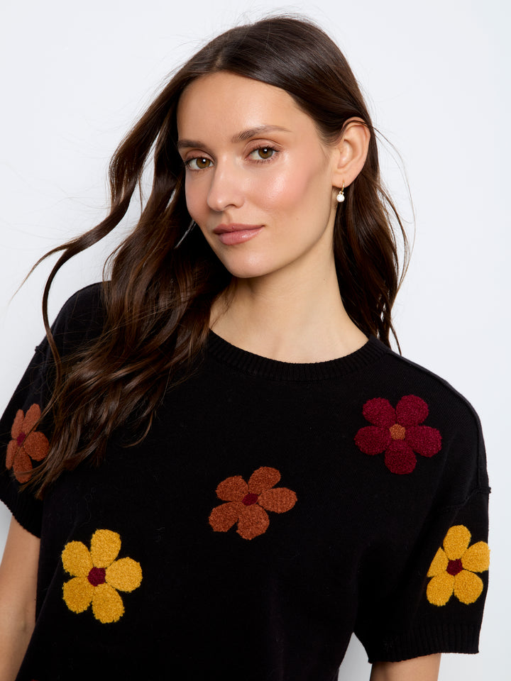 A woman wearing a black short-sleeve knit sweater with colorful 3D flower appliqués in yellow, brown, and burgundy, photographed against a clean white studio background.