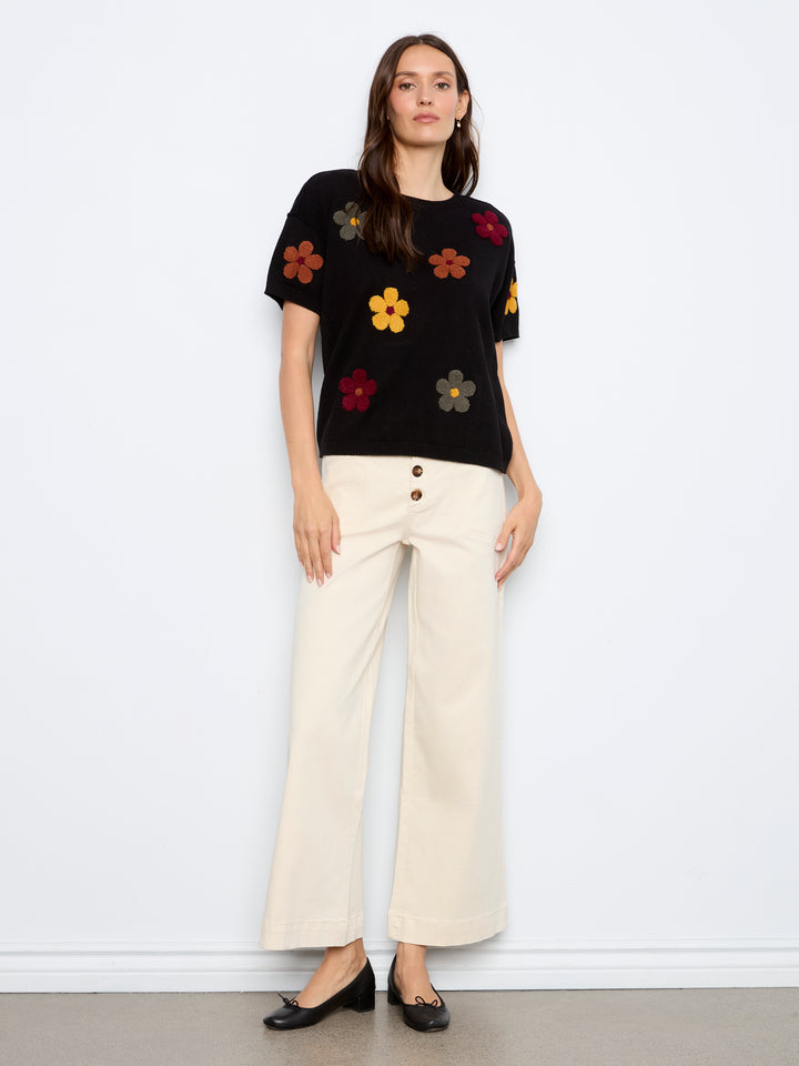 A woman stands against a plain white wall wearing a black short-sleeve knit top with colorful embroidered flowers, paired with cream wide-leg pants and black flats.