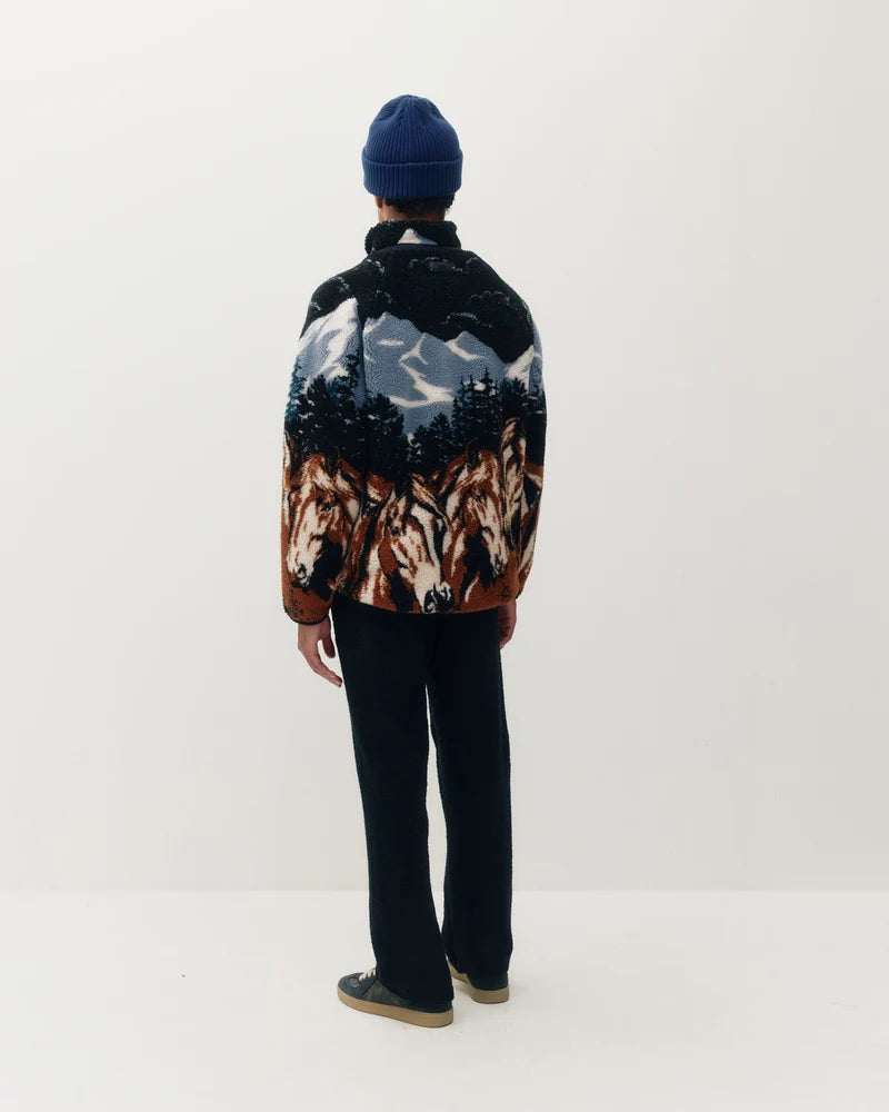 A person shown from the back wearing a dark knit sweater with a scenic mountain-and-horse print, black pants, and a blue beanie against a plain white studio background.