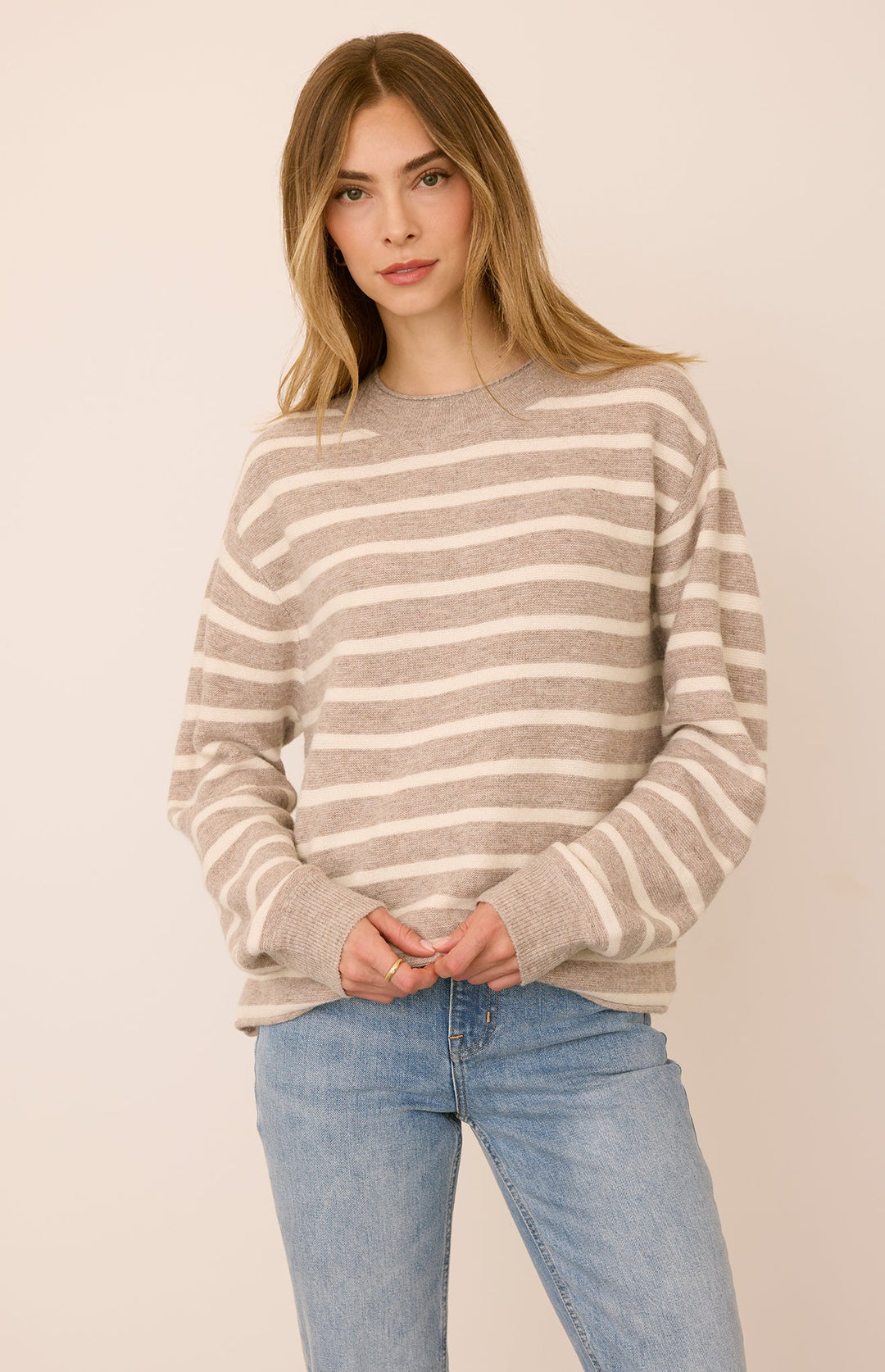 A woman wearing a beige and off-white horizontally striped crewneck sweater with light blue jeans, standing against a solid light beige background.