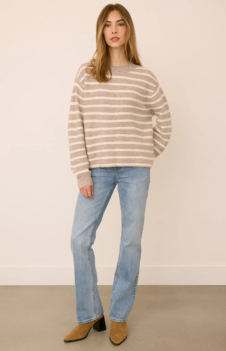A woman modeling a beige and white horizontally striped knit sweater, paired with light-wash blue jeans and tan ankle boots, standing against a plain cream wall.