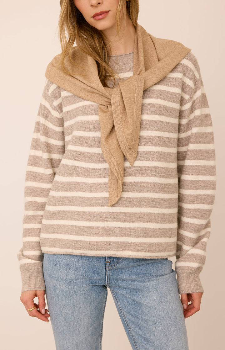 A woman wearing a beige and cream horizontally striped knit sweater with a tan knit scarf tied around the neck, paired with light blue jeans, against a solid light beige background.