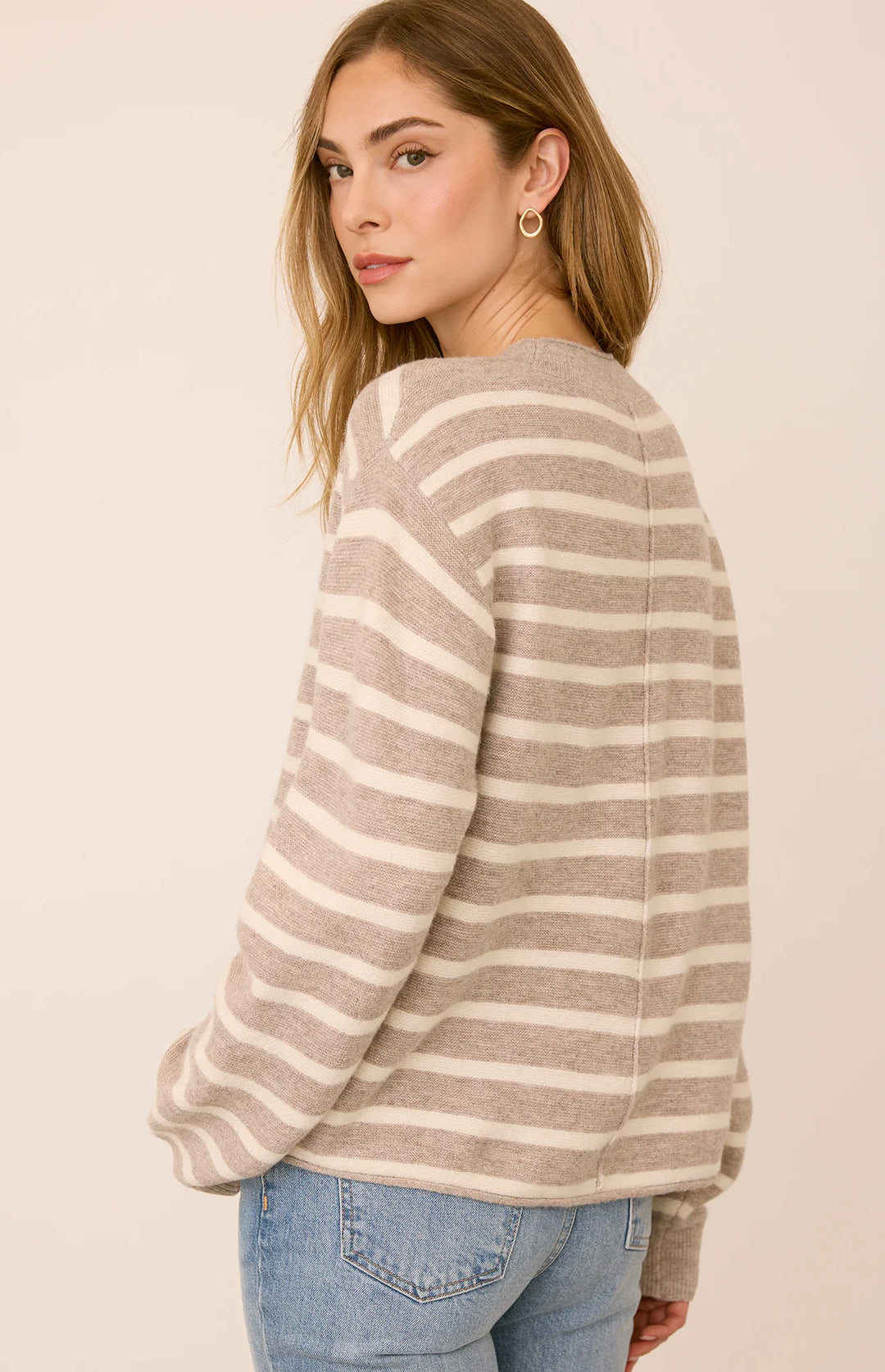 A woman wearing a beige and off-white horizontally striped knit sweater, shown from a three-quarters back view against a solid light beige background.