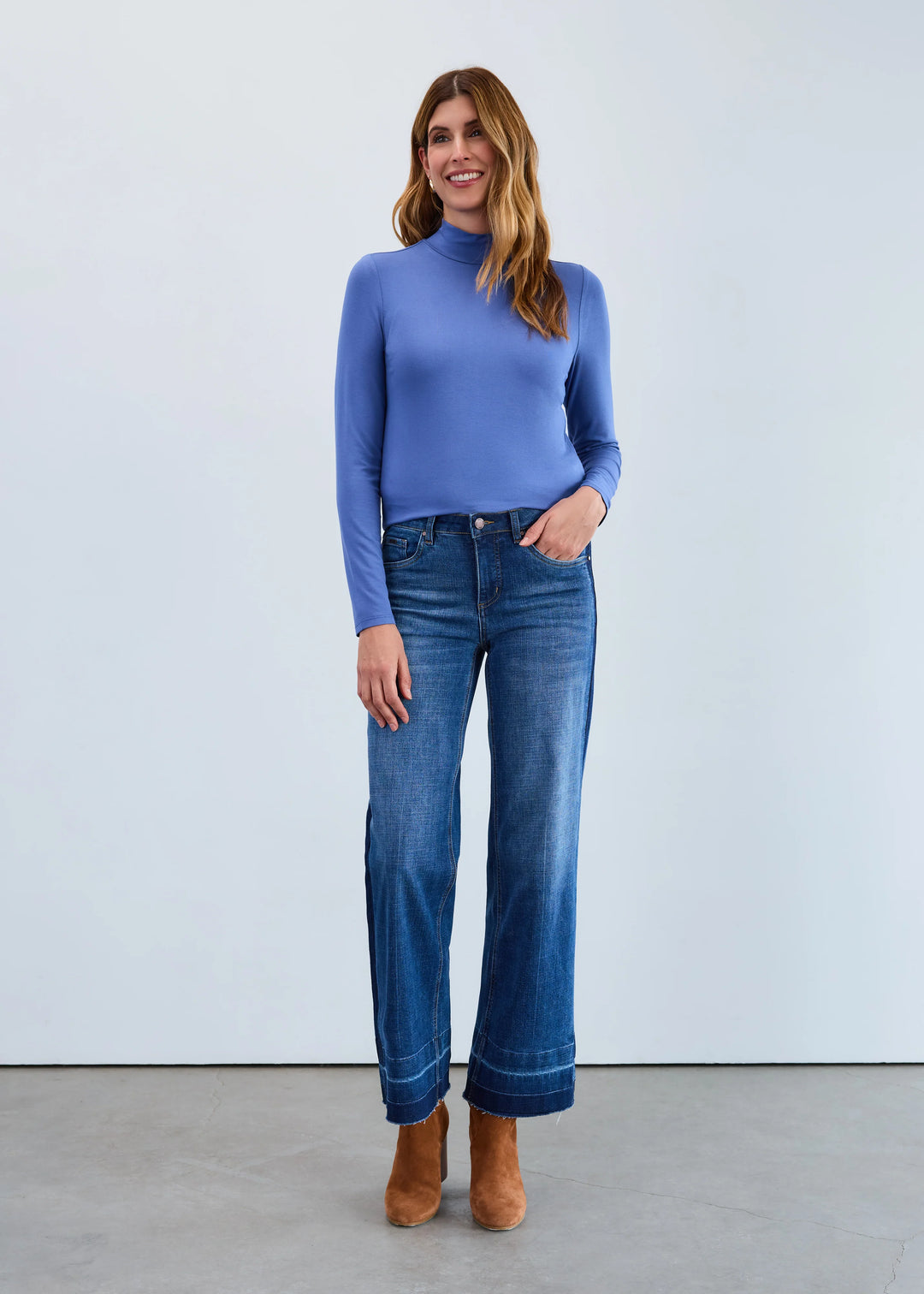 Woman wearing a blue turtleneck sweater and jeans standing against a light gray background