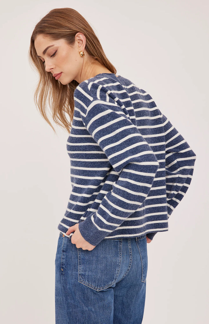 A woman wearing a navy blue and white horizontally striped long-sleeve knit top with voluminous balloon sleeves, paired with blue jeans, shown from a side-back angle against a light gray studio background.