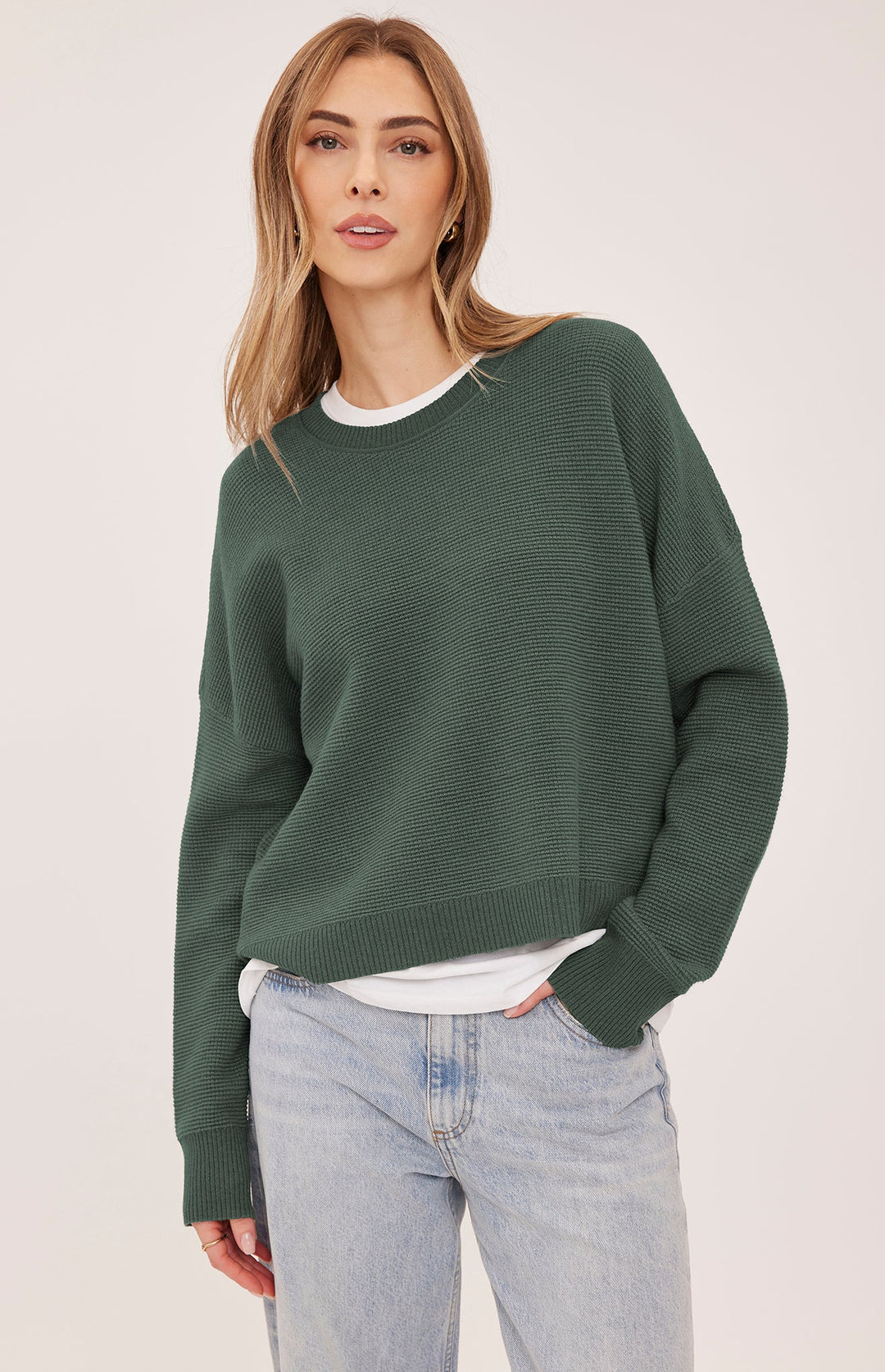 A woman wearing an oversized dark green ribbed knit sweater over a white t-shirt and light wash jeans, photographed against a plain light beige studio background.