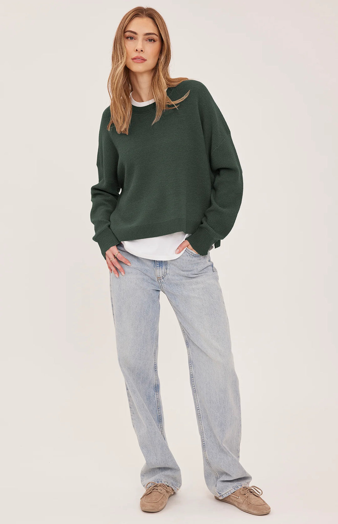 A woman modeling a dark forest green crewneck sweatshirt, styled with a white t-shirt, light-wash wide-leg jeans, and brown suede shoes, against a plain off-white studio background.