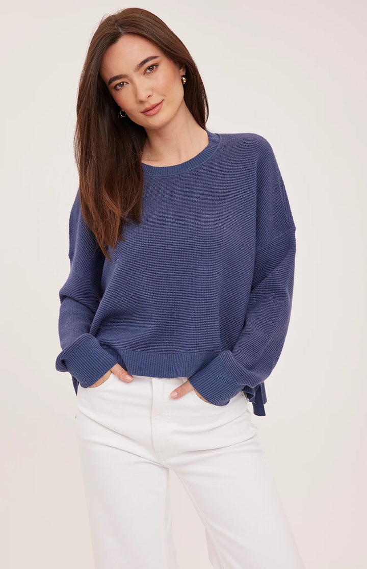 A woman wearing a ribbed knit crewneck sweater in a dusty periwinkle blue color, paired with white pants, against a plain light background.