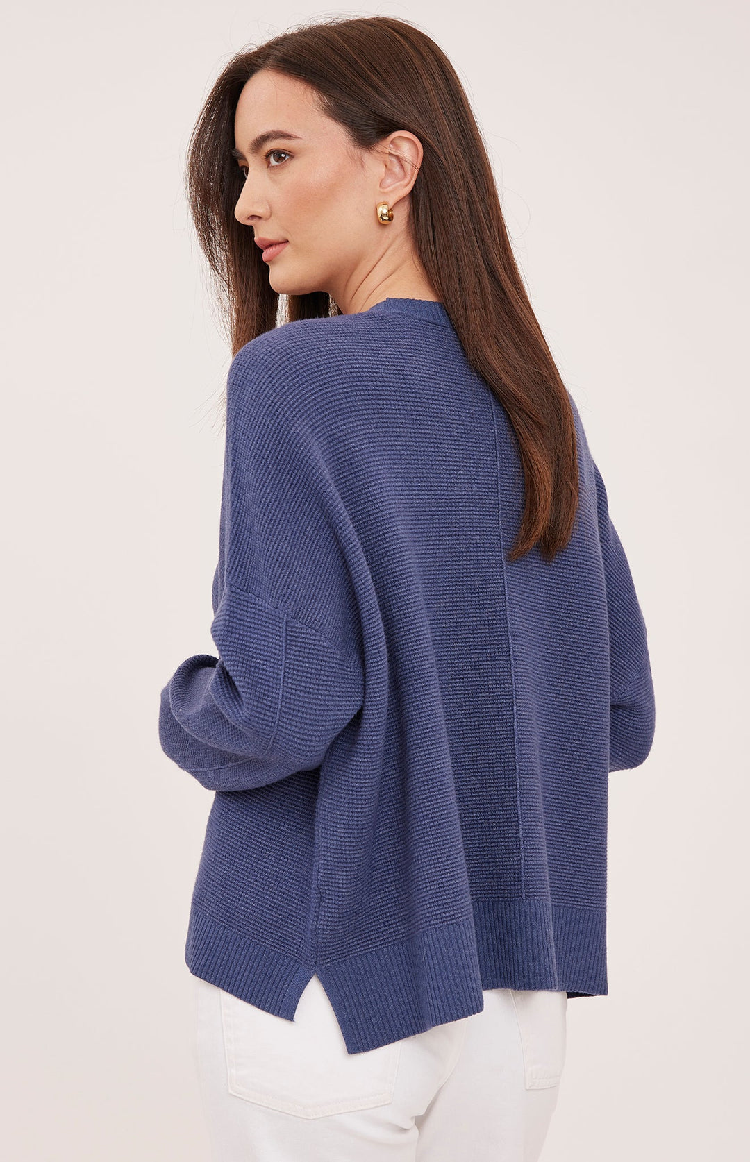 A woman wearing a textured, ribbed knit sweater in a dusty blue shade, shown from a three-quarter back view against a plain off-white studio background.