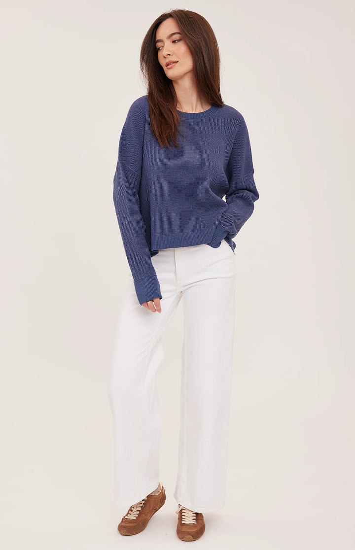 A woman modeling a blue crewneck knit sweater, white wide-leg trousers, and brown suede sneakers against a plain off-white studio background.