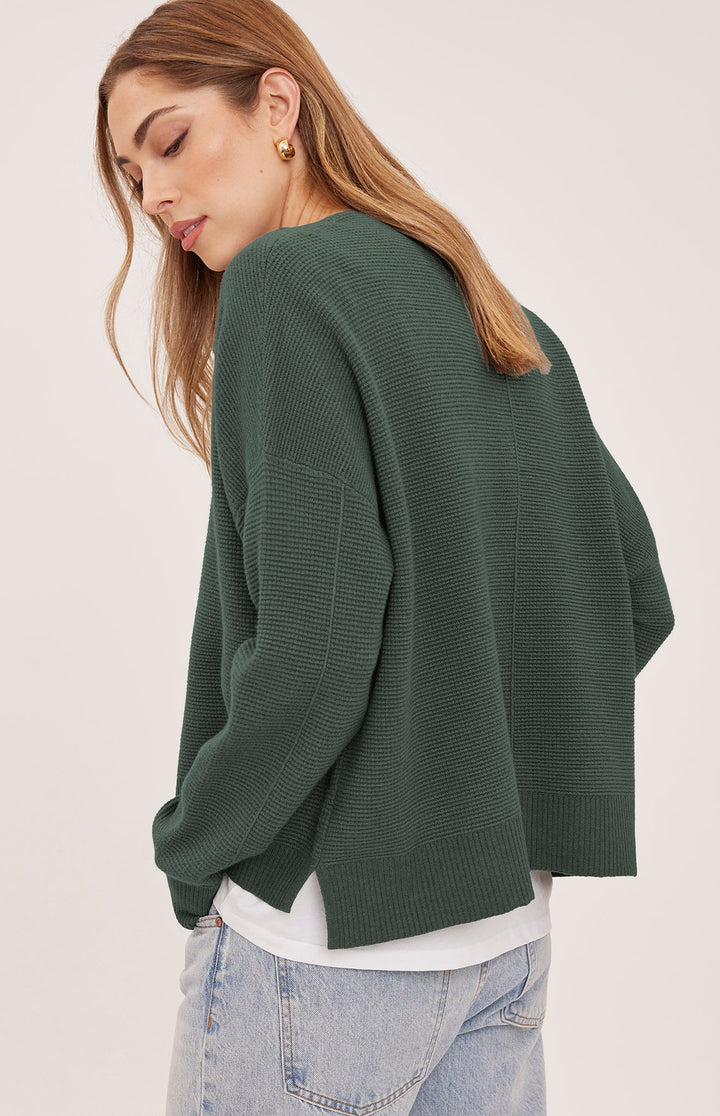 A woman wearing a dark green ribbed knit sweater, shown from a side-back angle against a plain light background.