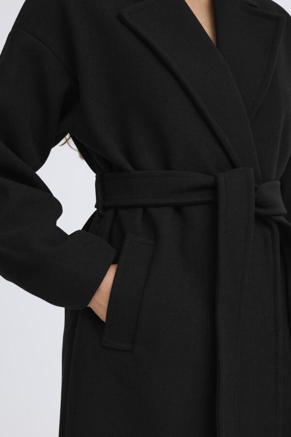 A close-up of a black wool coat with a tie belt, worn by a model whose hand is in a pocket, against a light gray background.