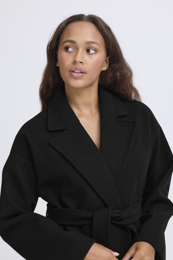 A woman wearing a black belted wrap coat or jacket, photographed against a plain light gray studio background.