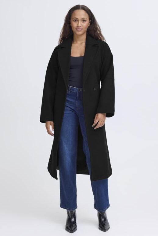 A woman modeling a long black coat over a dark top and blue jeans, standing against a plain light gray studio background.