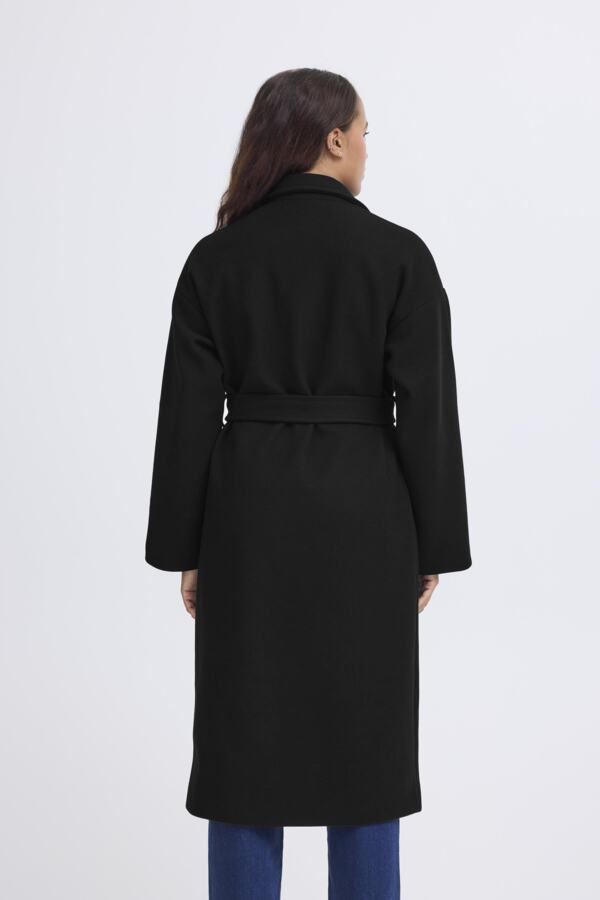 A woman viewed from the back wearing a long black belted coat over blue jeans, photographed against a plain light gray studio background.