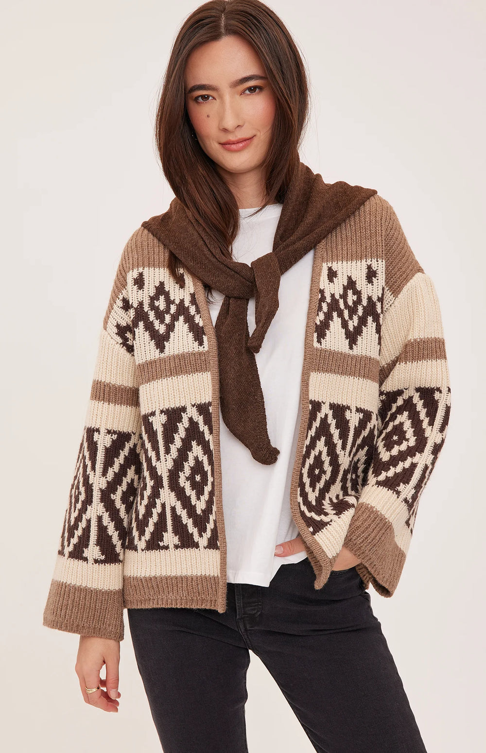 A woman models a brown and cream knit cardigan with a hood and a matching brown scarf tied around the neck, worn over a white t-shirt and black jeans against a light beige studio background.