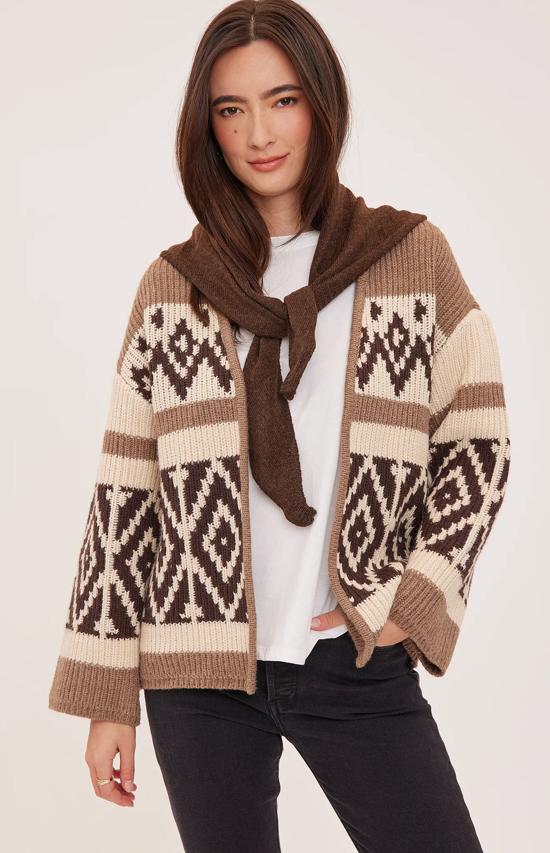A woman models a brown and cream knit cardigan with a hood and a matching brown scarf tied around the neck, worn over a white t-shirt and black jeans against a light beige studio background.