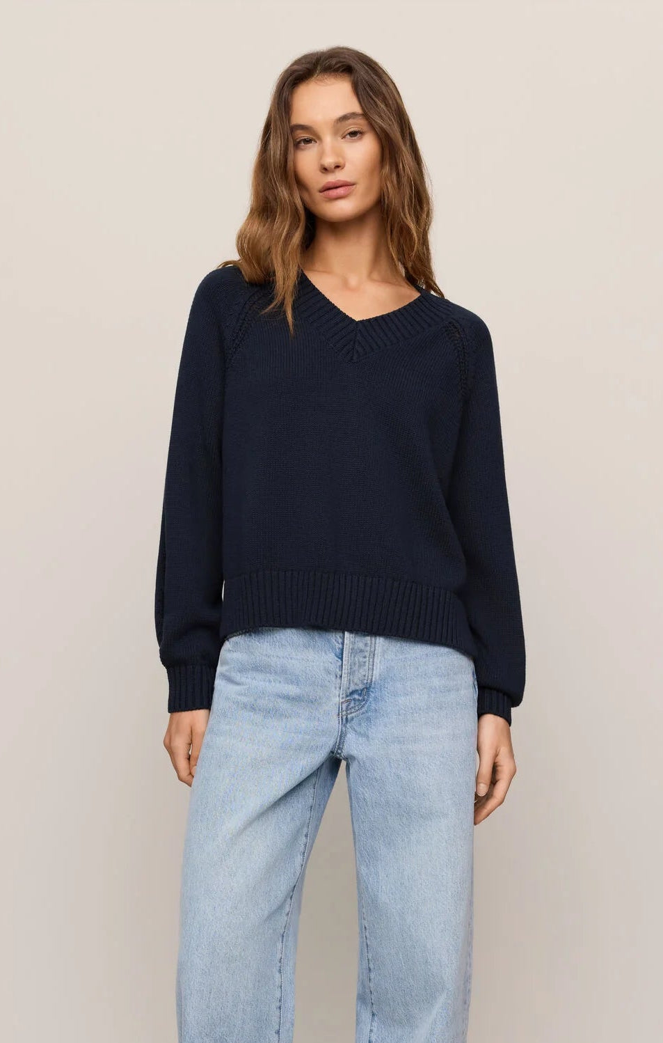 A woman modeling a dark navy blue V-neck knit sweater and light-wash wide-leg jeans against a plain beige studio background.