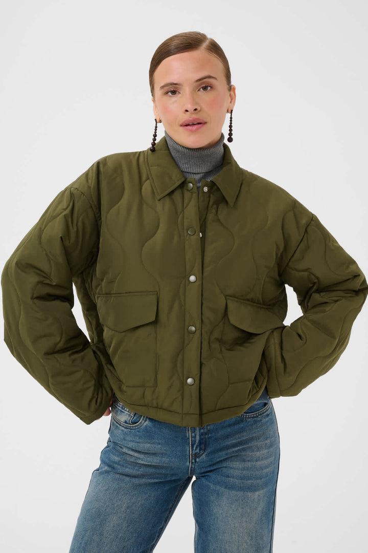 A woman modeling an olive green quilted puffer jacket with a button-up front and flap pockets, styled over a gray turtleneck and blue jeans against a light gray studio background.