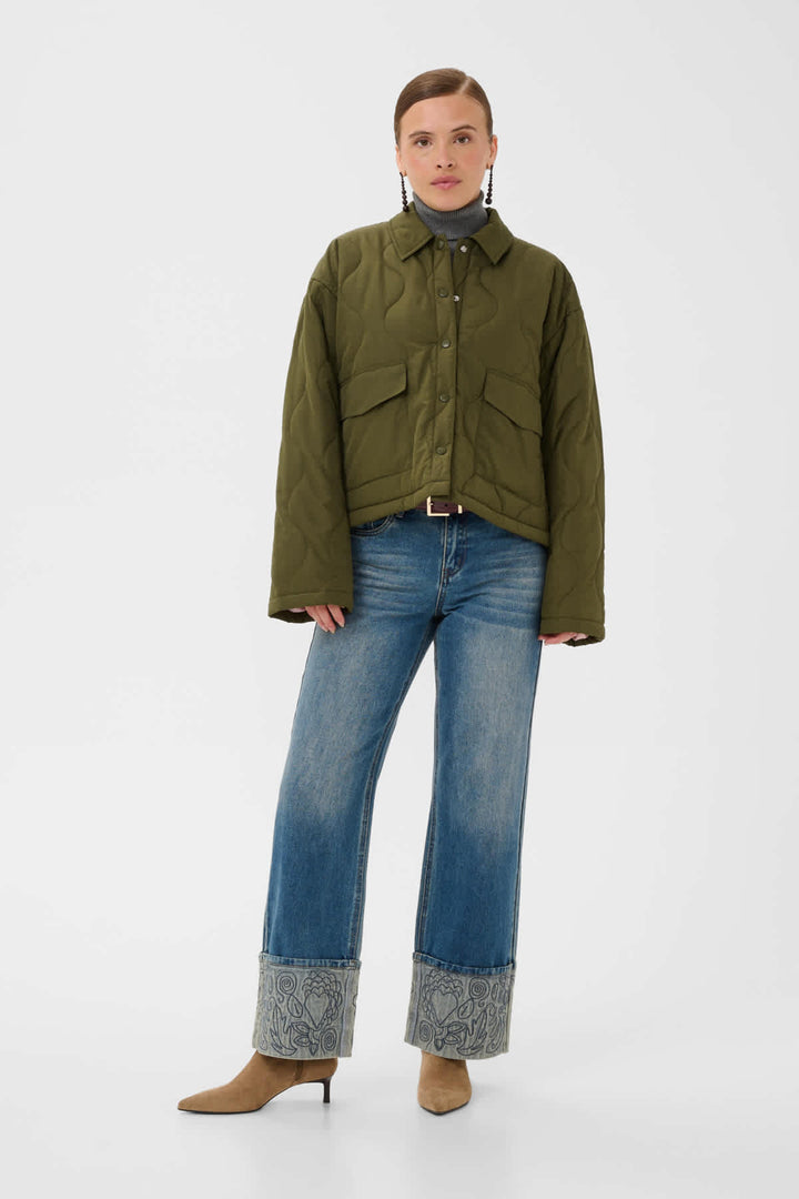 A model stands against a light gray studio background wearing an olive green quilted jacket, blue jeans with patterned cuffed hems, and tan pointed-toe heels.