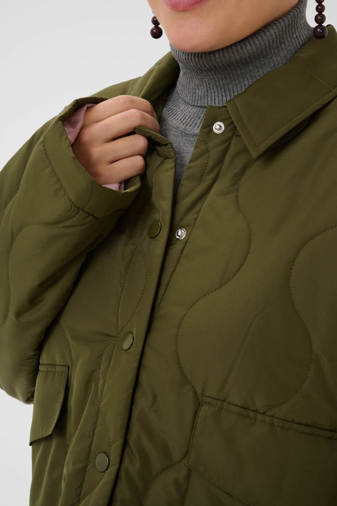 Close-up of a person wearing an olive green quilted jacket over a gray ribbed turtleneck sweater, with a hand adjusting the collar and buttons.