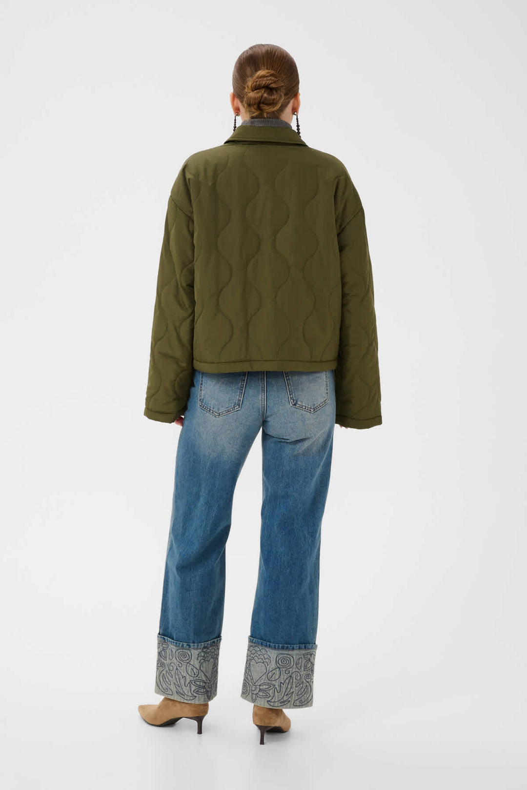 A model viewed from the back wearing an olive green quilted jacket, blue jeans with patterned cuffs, and tan heels, set against a plain light gray studio background.