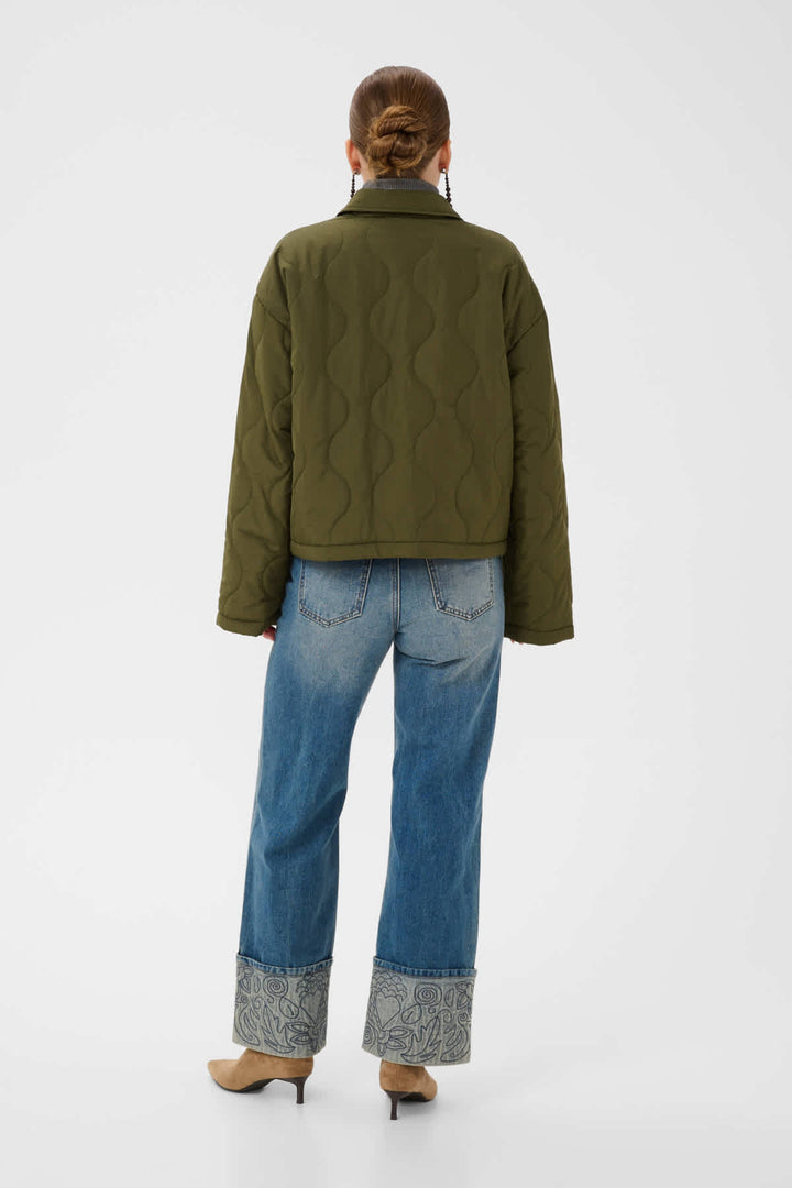 A model viewed from the back wearing an olive green quilted jacket, blue jeans with patterned cuffs, and tan heels, set against a plain light gray studio background.
