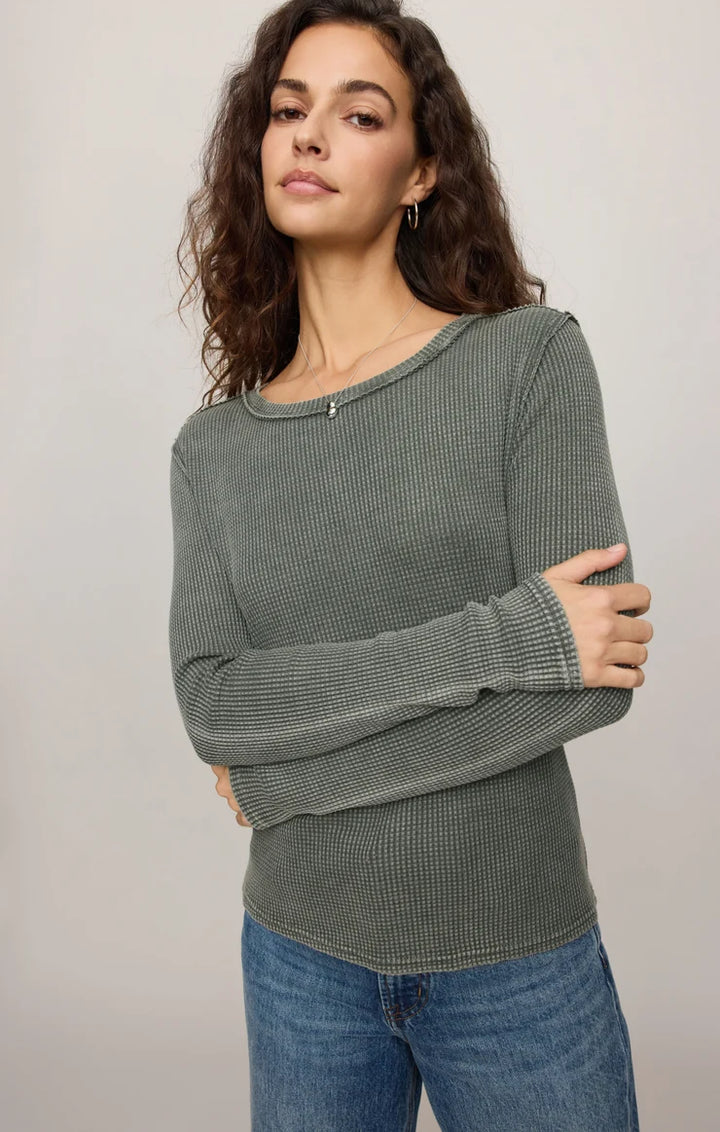 A woman wearing a ribbed long-sleeve knit top in a muted sage green color, paired with blue jeans, photographed against a plain light gray studio background.
