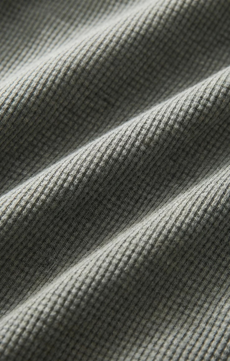 Close-up of a waffle-knit fabric in a muted gray-green color, showing the textured knit pattern and soft folds.
