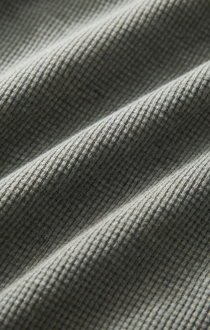 Close-up of a waffle-knit fabric in a muted gray-green color, showing the textured knit pattern and soft folds.