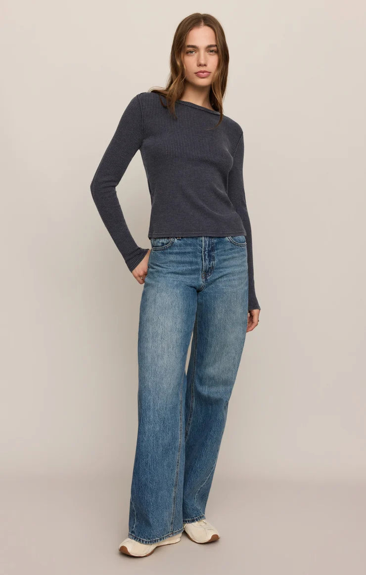 A woman modeling a dark charcoal gray long-sleeve knit top with an asymmetrical neckline, paired with wide-leg blue jeans and beige shoes, against a solid light beige studio background.